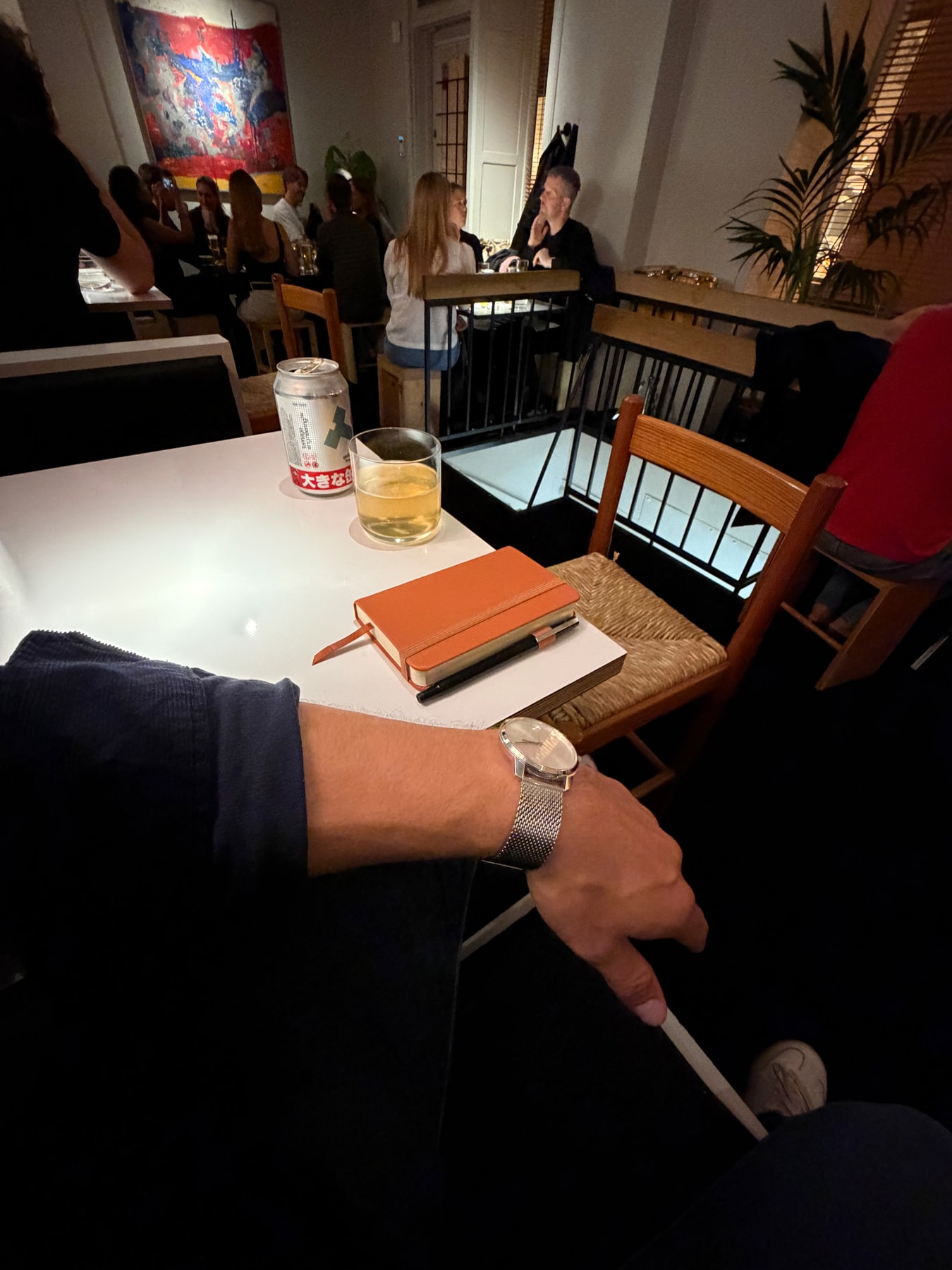 An orange notebook and drink on a restaurant table, with a watch-wearing wrist in the foreground.