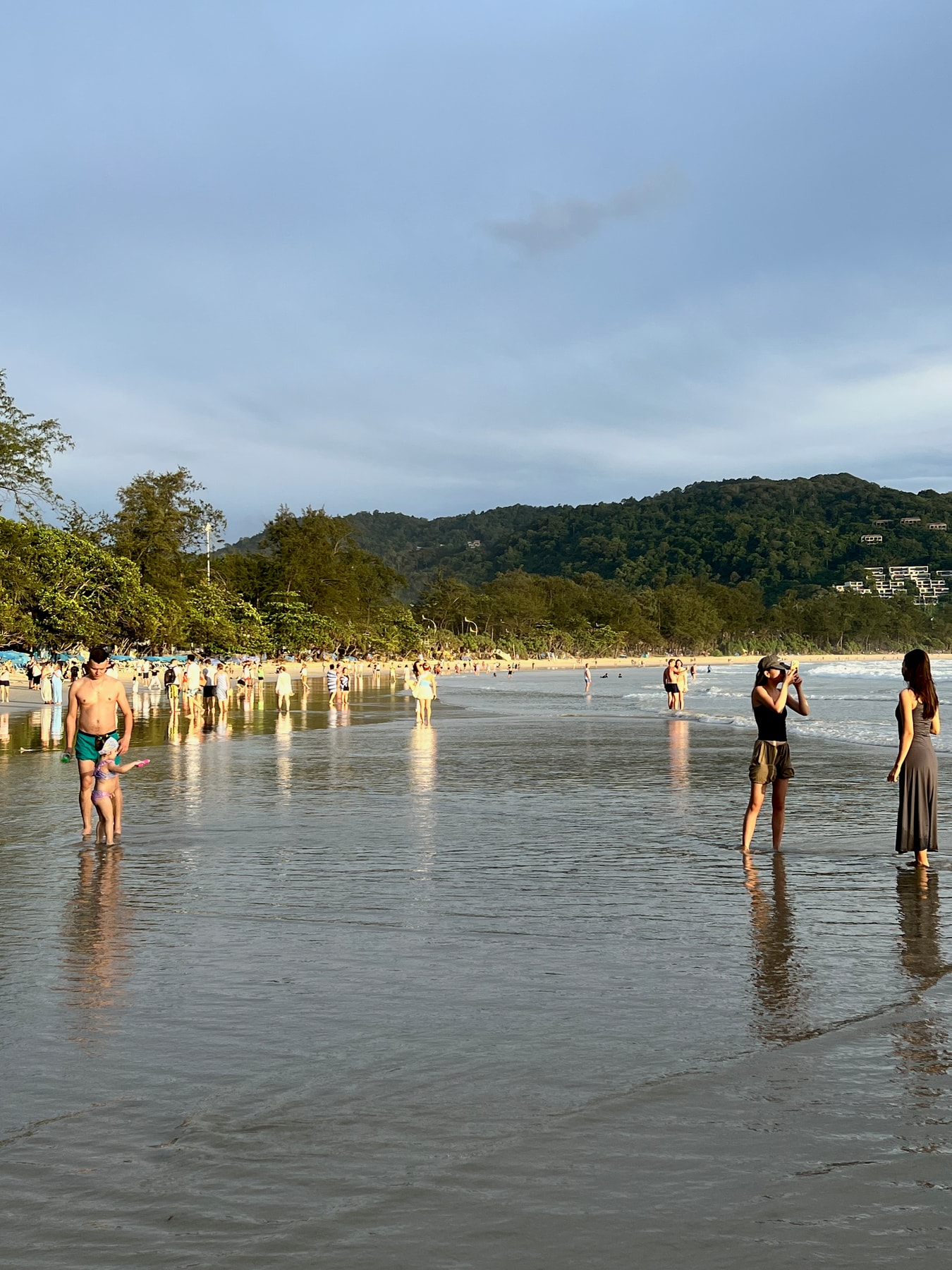 People stand and walk in the shallow water of a beach in Thailand at sunset.
