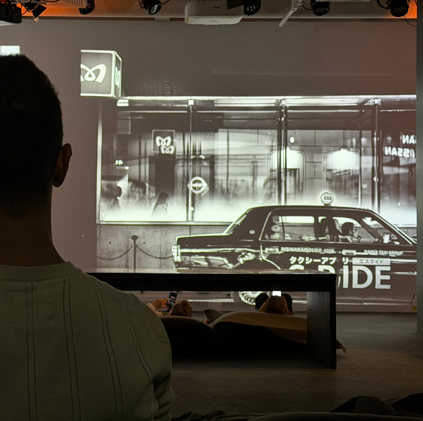 A person seated before a large projection of a Tokyo street and taxi, with other visitors reflected below.